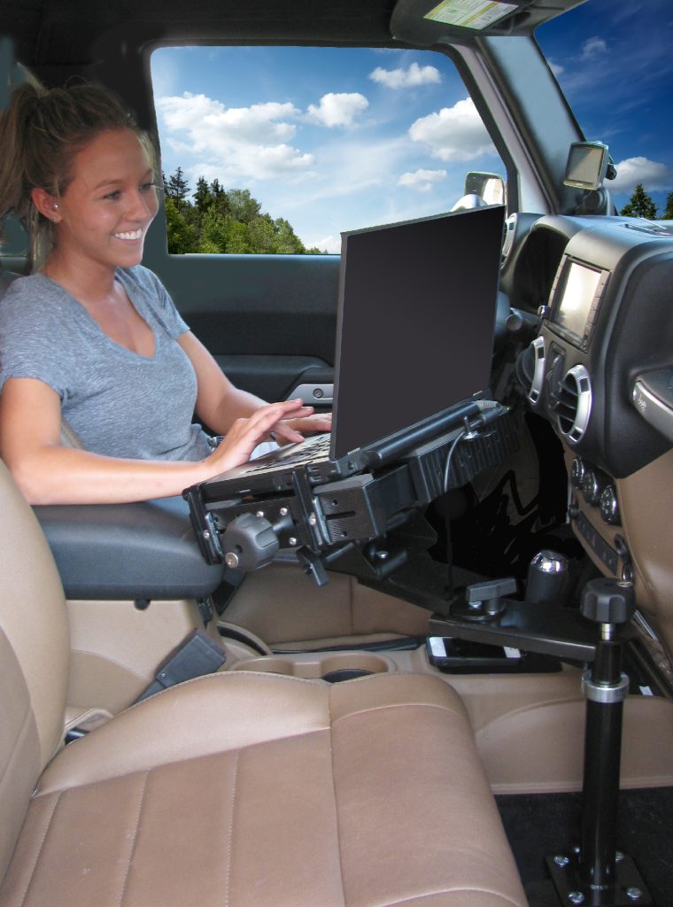 A woman types on a laptop mounted to a stand inside a car's front passenger seat area.